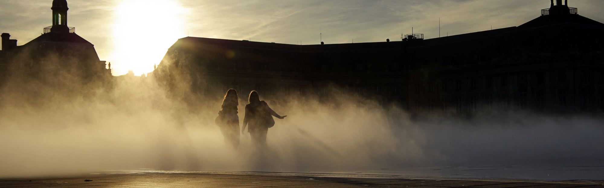 The place de la Bourse in Bordeaux at sunset, its long classical frontage in silhouette above the flooded stone of the water mirror, with mist drifting across it from the fountains.