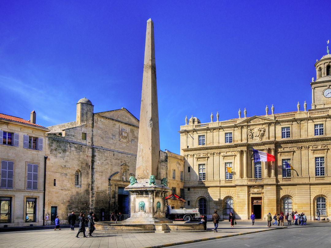 The obelisk on the place de la République at Arles, with the town hall and the Romanesque church front behind it under a hard blue sky.