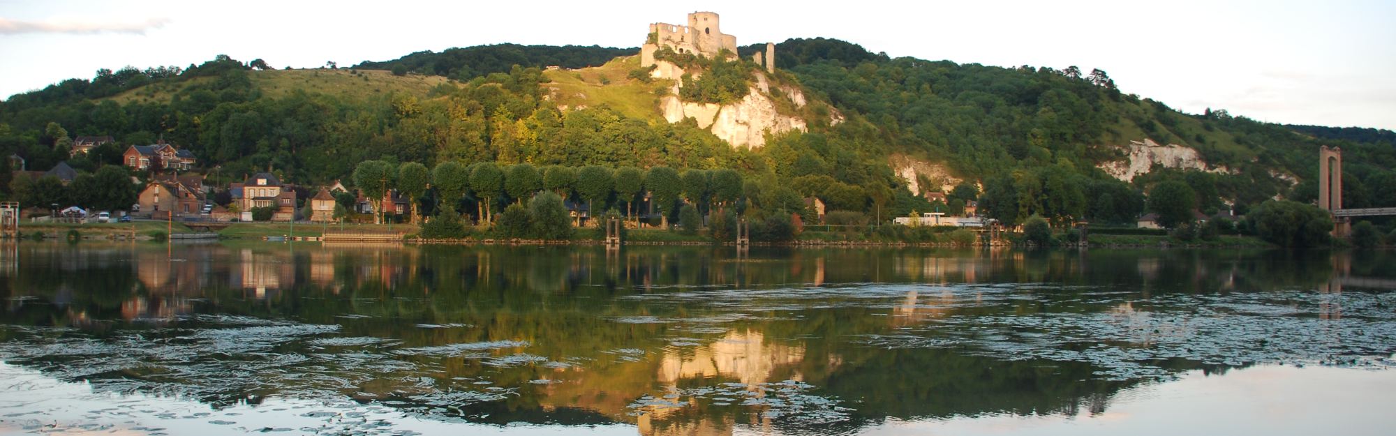 Château Gaillard standing on a wooded chalk ridge above the Seine at Les Andelys, the ruin and the slope reflected in still water at the end of the day.